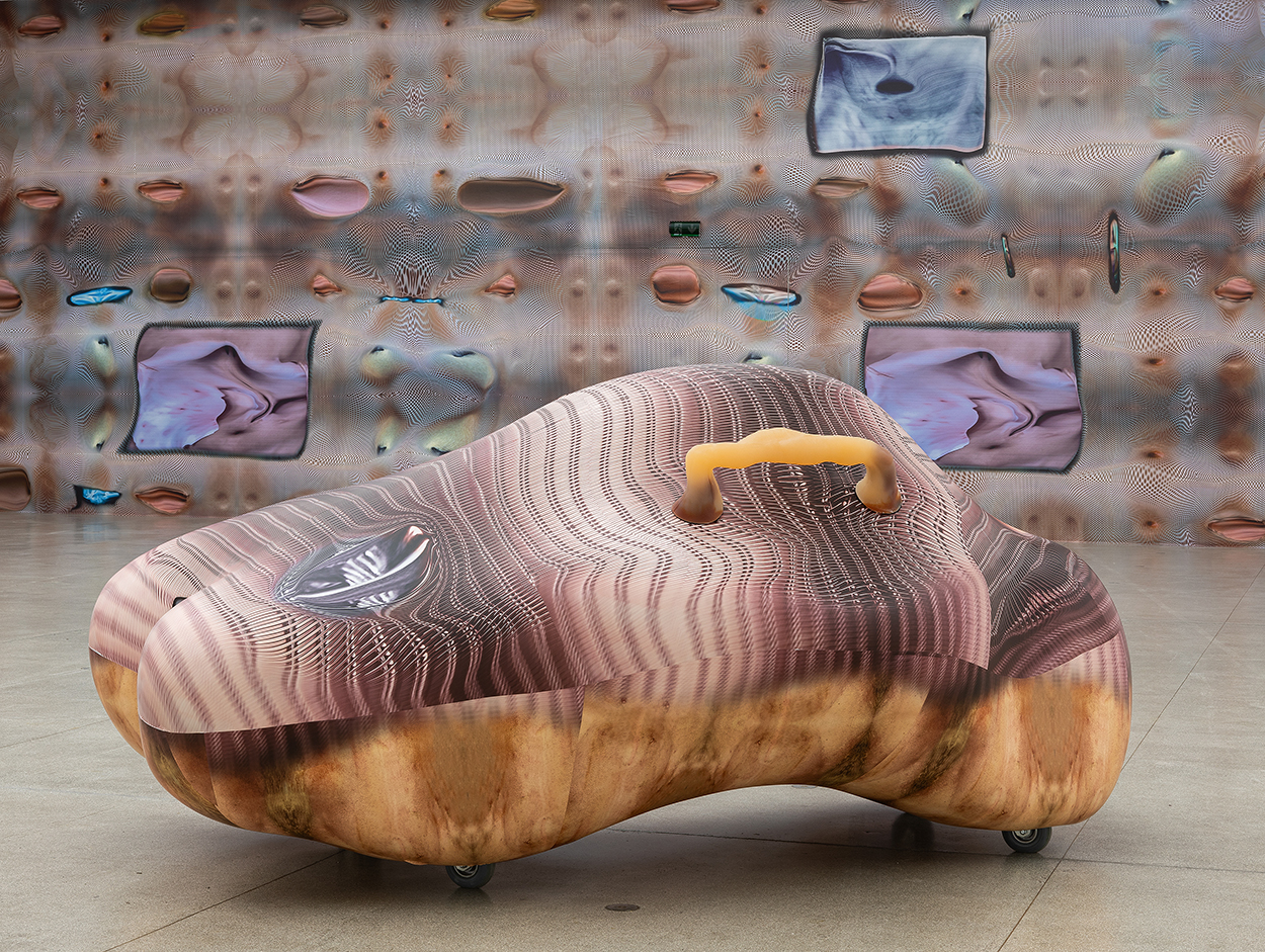 An abstract, lumpy, supine sculpture has a flesh-like grab bar. It sits in front a wall covered in ambiguous orifices.