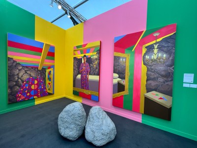 A brightly hued fair booth with three paintings, one of which shows a bathroom whose walls are painted in similar colors. At the center are two sculptures resembling rocks.