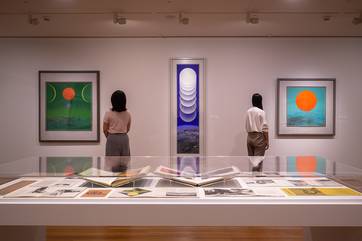 Three colorful paintings of hovering orbs hang on a wall in front of a vitrine.