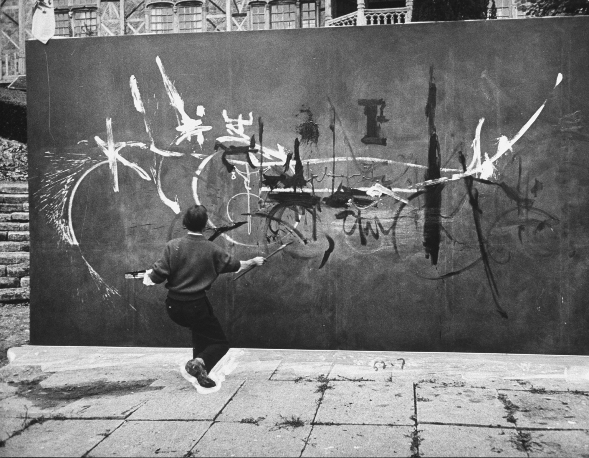 A photograph captures Georges Mathieu in the midst of painting a large abstract canvas outside while wielding long brushes.