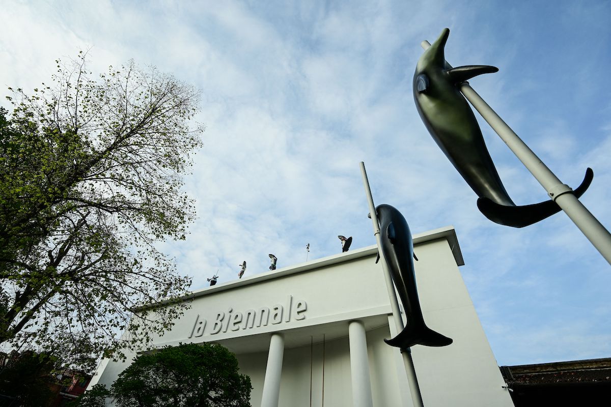 A building whose columned facade reads 'la Biennale.' Running across its top and in front of it are sculptures of dolphins attached to poles.