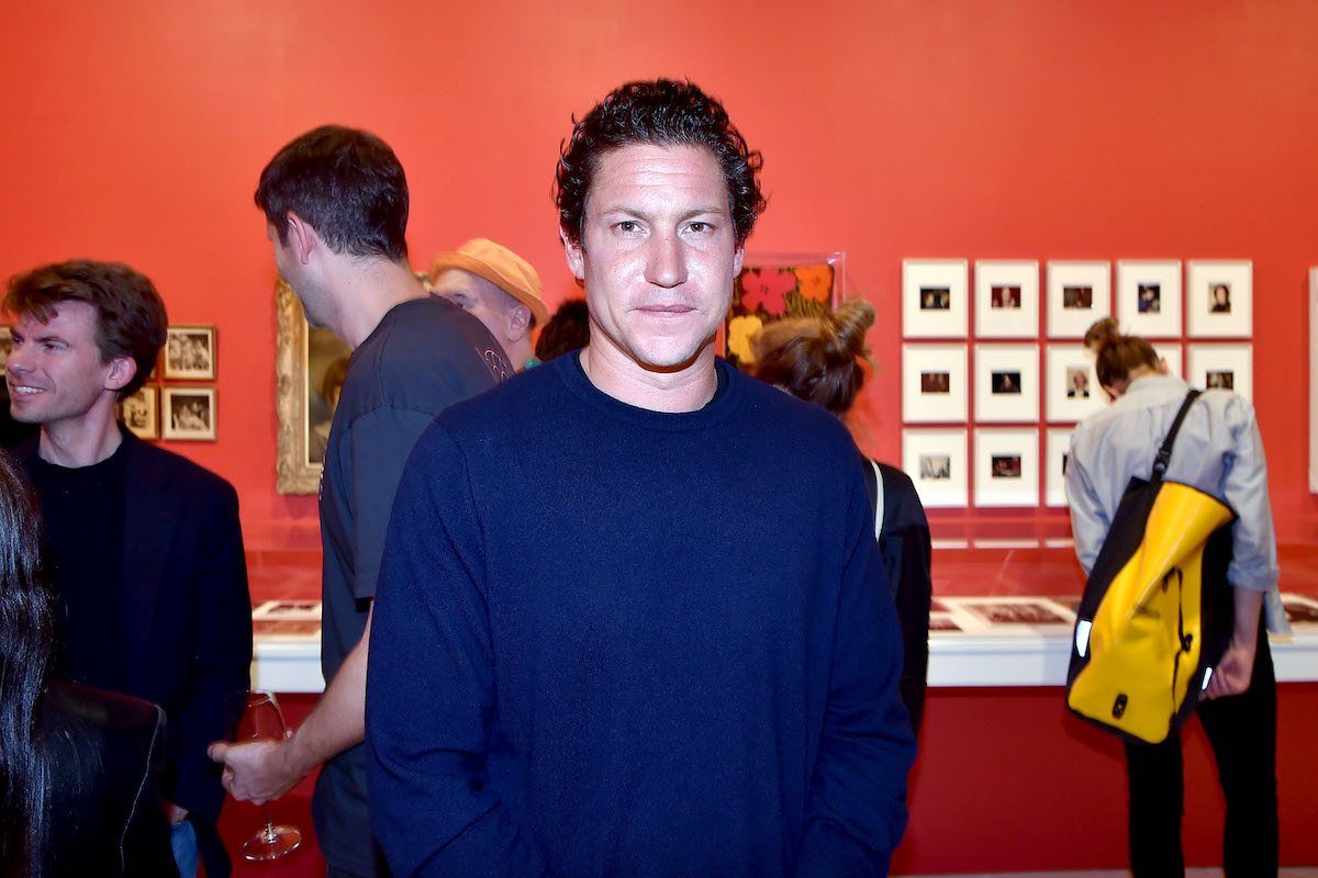 A white man in a blue sweater standing in a gallery with other people looking at photographs around him.
