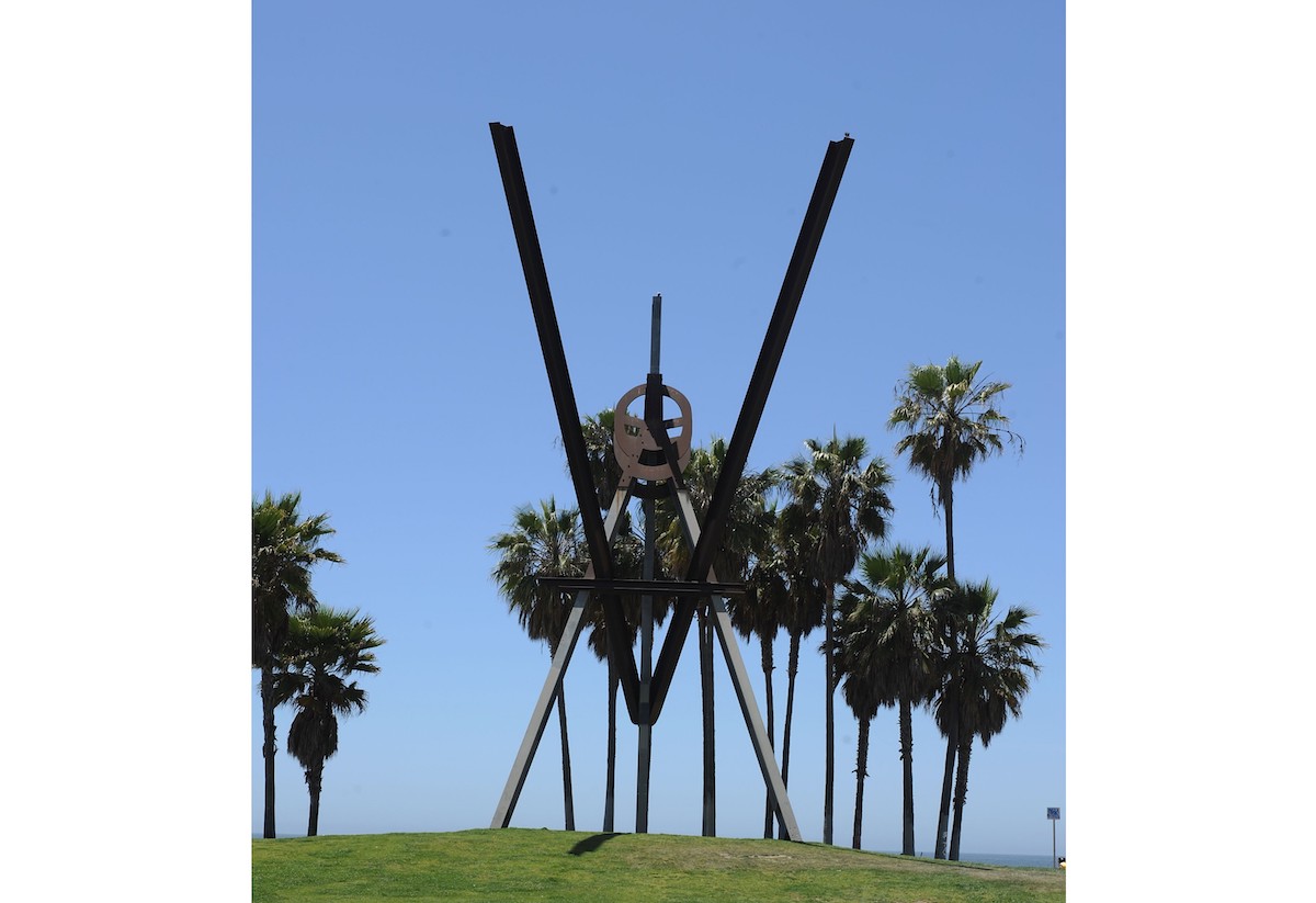 A giant steel sculpture composed of beams arranged in V-like forms. The sculpture is set on a grassy hill with palm trees.
