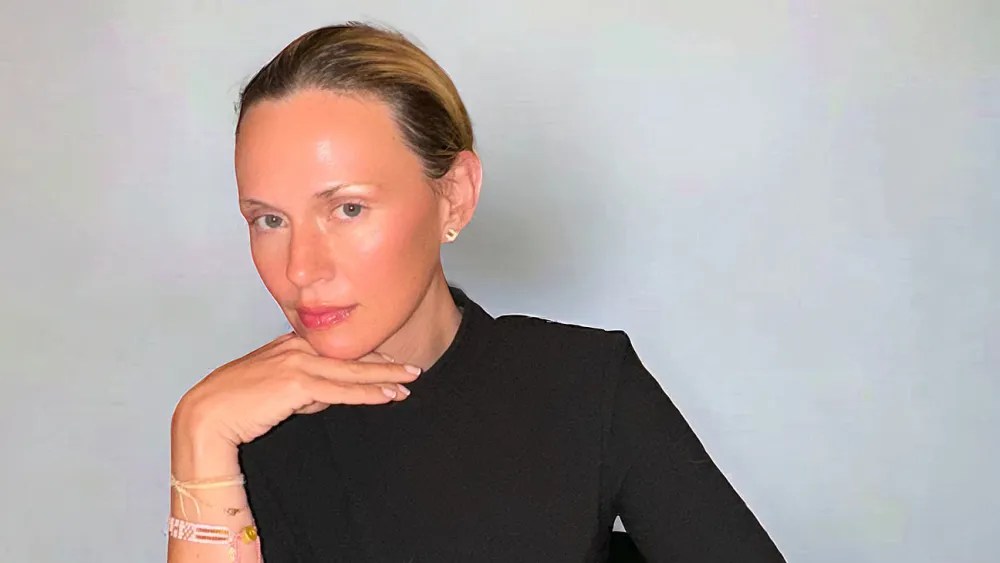 A white woman with dark blonde hair wearing a black long-sleeve top, looks at the camera.