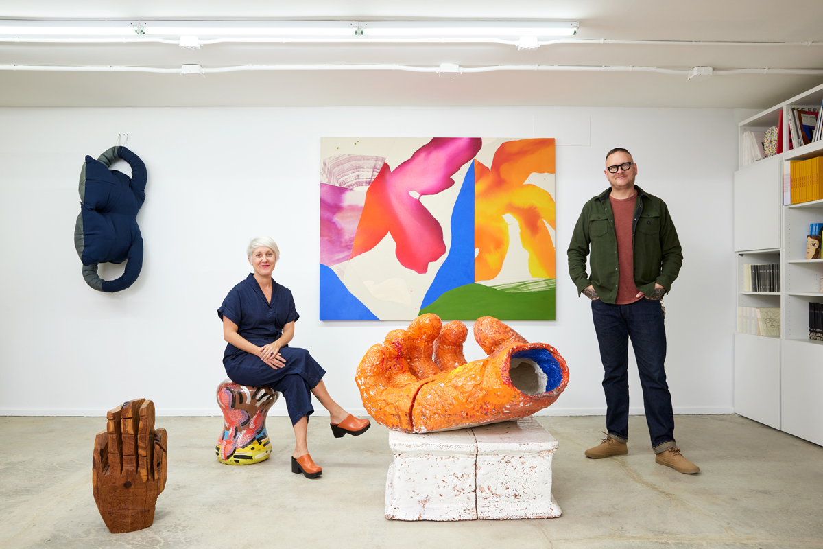 A white woman sits on a stool that is an artwork and a man stand to her right. They pose in front of several artworks in an exhibition.