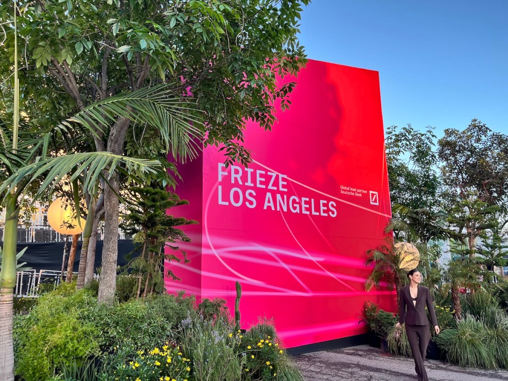 A woman in a blazer posing before a gigantic pink sign near palm trees that reads 'FRIEZE LOS ANGELES.'