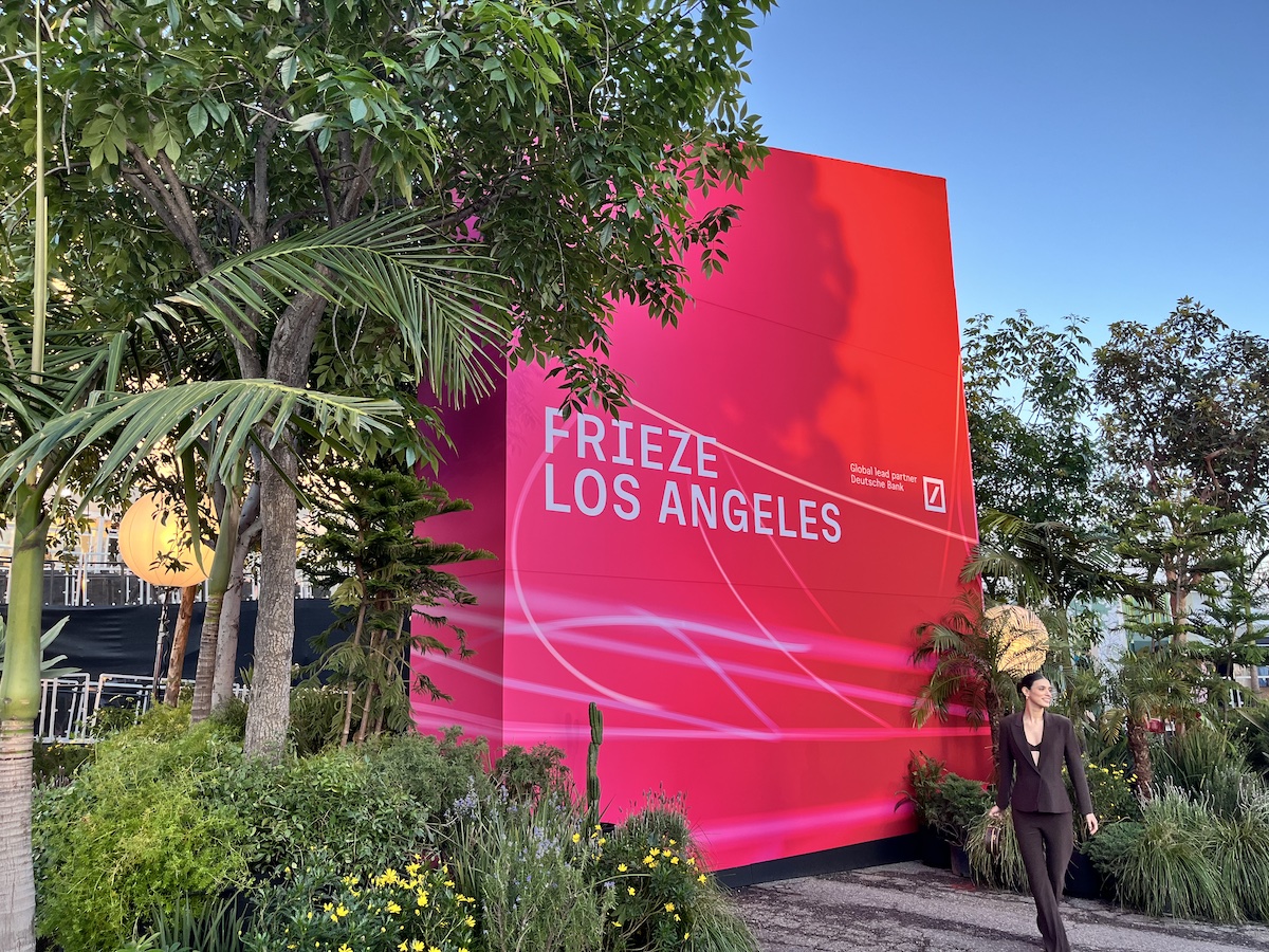 A woman in a blazer posing before a gigantic pink sign near palm trees that reads 'FRIEZE LOS ANGELES.'