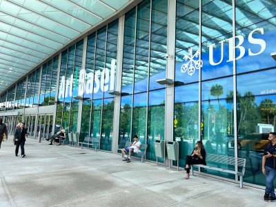 A convention center with the words Art Basel on its windows. Several people sit on benches in front of it.