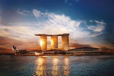 Exterior view of a building seen from across a harbor. There are several low-lying buildings in front of three tilting, soaring towers that gleam yellow in the sunset. The towers are connected by one long outdoor platform that sits atop them.