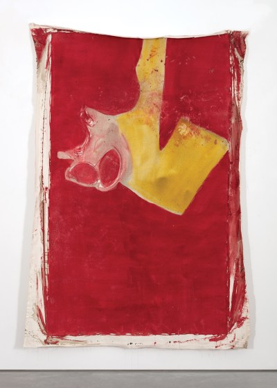 An unstretched canvas affixed to the wall. The canvas is mostly red with a swath of yellow and white in the upper section.