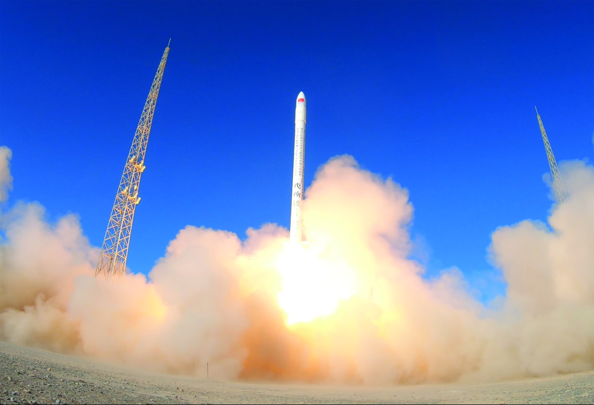 A fish eye photo of a long skinny rocket taking off into a blue sky. Clouds of dust are below.